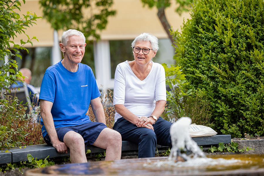 Ein Patient und eine Patientin sitzen auf einer Bank an einem Springbrunnen und lächeln in die Kamera.